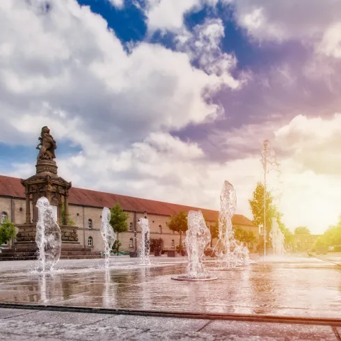 Ein zentraler Platz mit einem Brunnen, aus dem Wasser spritzt. Im Hintergrund sind historische Gebäude und ein strahlender Himmel zu sehen.