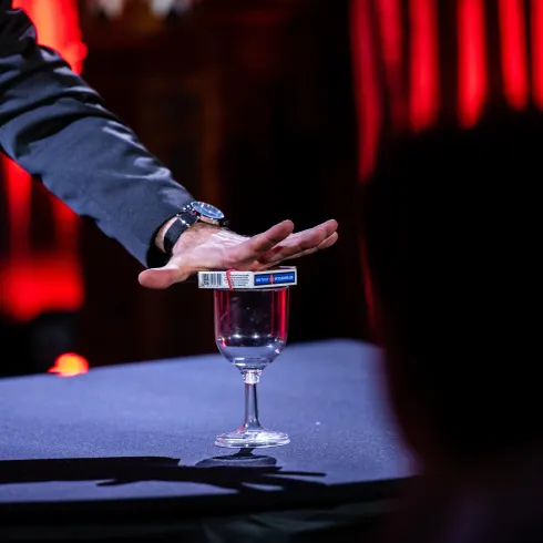 Ein Magier führt einen Trick vor, indem er eine Schachtel über einem Glas Wasser balanciert. Im Hintergrund sind rote Lichtakzente zu sehen.