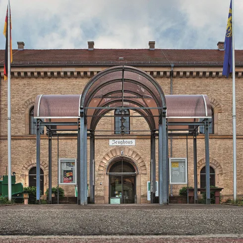 Ein beeindruckendes Gebäude mit einem modernen Eingangsbereich. Die Fassade besteht aus hellen Ziegeln und es sind Fahnen im Vordergrund zu sehen.