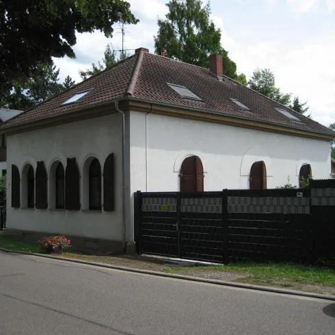 Ein zweigeschossiges Haus mit einem roten Ziegeldach und weißen Wänden. Es hat mehrere Fenster mit Fensterläden und ist von einem schwarzen Zaun umgeben.