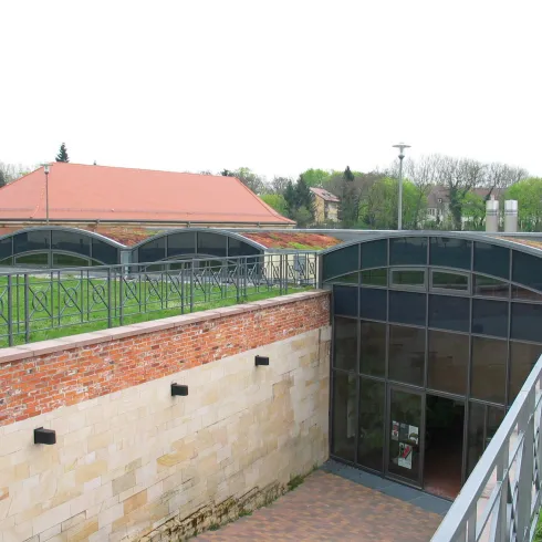 Ein modernes Gebäude mit einer geschwungenen Dachkonstruktion und großen Fenstern. Es liegt in einer grünen Umgebung mit Rasenflächen und Bäumen.