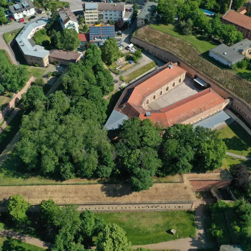 Eine alte Festung umgeben von Bäumen und Grünflächen. Im Hintergrund sind moderne Gebäude sichtbar.