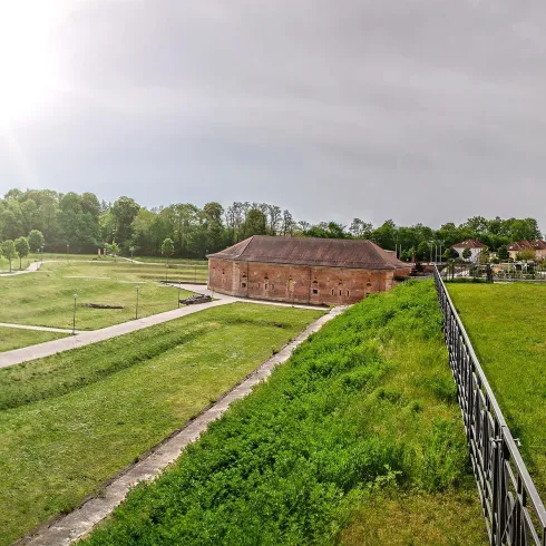 Eine weite Parklandschaft mit grünen Wiesen und Bauten im Hintergrund. Der Himmel ist teilweise bewölkt und es strahlt Licht durch.