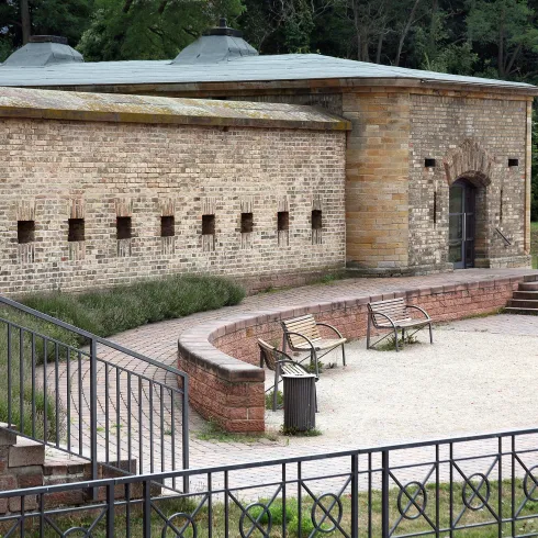 Ein altes, steinernes Gebäude mit Fenstern und einer Terrasse. Im Vordergrund sind Bänke und eine bepflanzte Umgebung zu sehen.