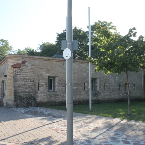 Ein historisches Gebäude aus Stein umgeben von Bäumen und Gras. Im Vordergrund ist ein Gehweg mit einem Straßenlampe sichtbar.