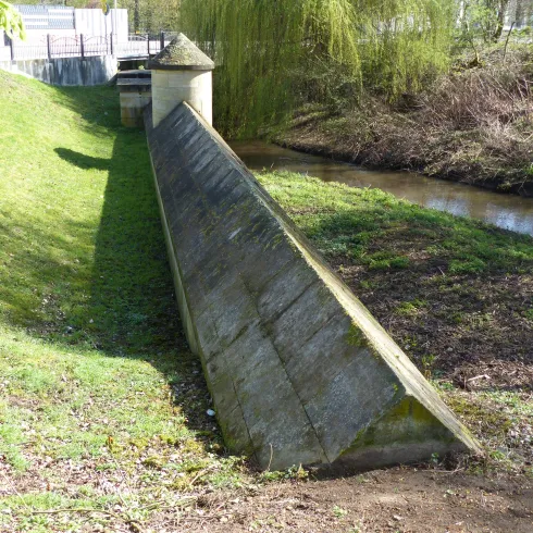 Ein schmaler betonierter Kanal mit einer geneigten Mauer an einem kleinen Fluss. Umgeben von grünem Gras und Bäumen im Hintergrund.