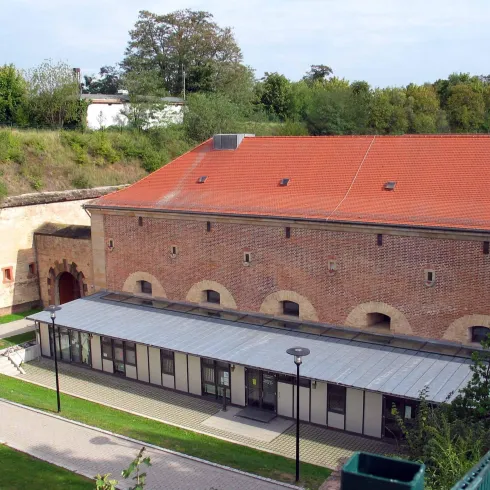 Eine alte Mauer mit einem roten Dach und einem modernen Anbau aus Glas. Umgeben von Grünflächen und Bäumen.