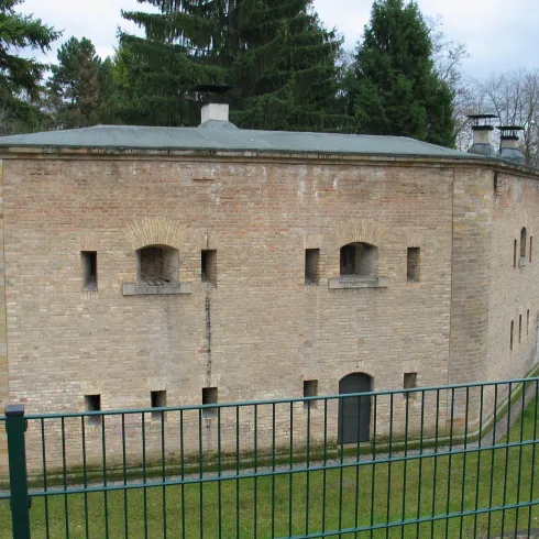 Ein historisches Gebäude aus Backstein mit Fenstern und einem Flachdach. Es ist von einem grünen Bereich umgeben und hat einen Zaun im Vordergrund.