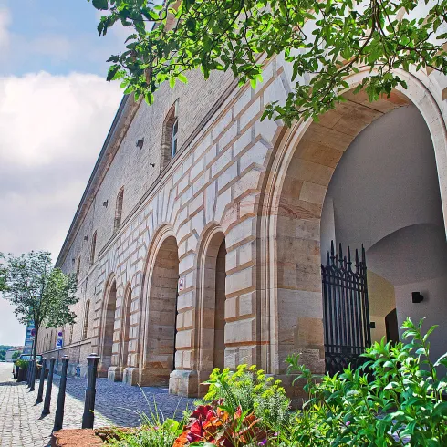 Ein historisches Gebäude mit einer schönen Steinfassade und mehrere Bögen. Im Vordergrund sind Pflanzen und Bäume zu sehen, die das Bild lebendiger machen.