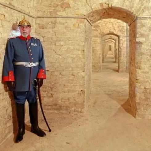 Ein historischer Soldat steht in einem Steinbögen-Korridor. Die Wände sind aus grobem Stein, und der Raum wirkt vielseitig und geheimnisvoll.