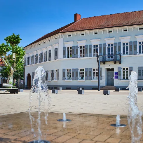 Ein schönes Gebäude mit einem klaren blauen Himmel und einem Springbrunnen im Vordergrund. Die Umgebung ist einladend mit Bäumen und Sitzgelegenheiten.