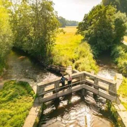 Eine Holzbrücke über einen kleinen Fluss, umgeben von grünen Bäumen und Wiesen. Im Hintergrund sind weitere Bäume und eine ländliche Landschaft zu sehen.