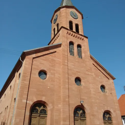 Eine beeindruckende Kirche aus rotem Stein unter einem klaren blauen Himmel. Der Kirchturm ist mit einer Uhr und einem Wetterfahne geschmückt.