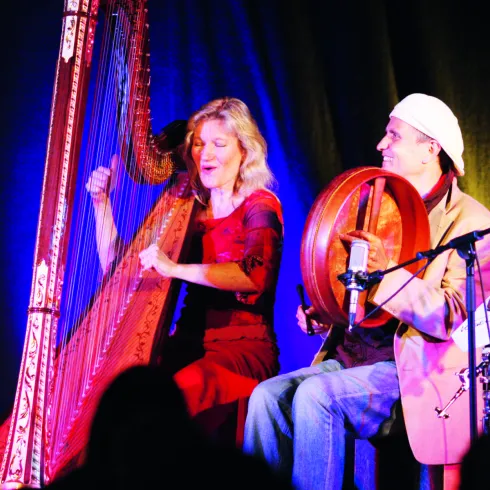 Zwei Musiker auf der Bühne, eine Frau spielt Harfe und ein Mann ein Handtrommeln. Die Atmosphäre ist lebhaft und musikalisch.