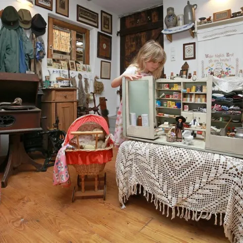 Ein kleines Mädchen erkundet einen nostalgischen Raum voller antiker Möbel und Dekorationsobjekte. Auf einem Tisch steht eine detailreiche Puppenküche.