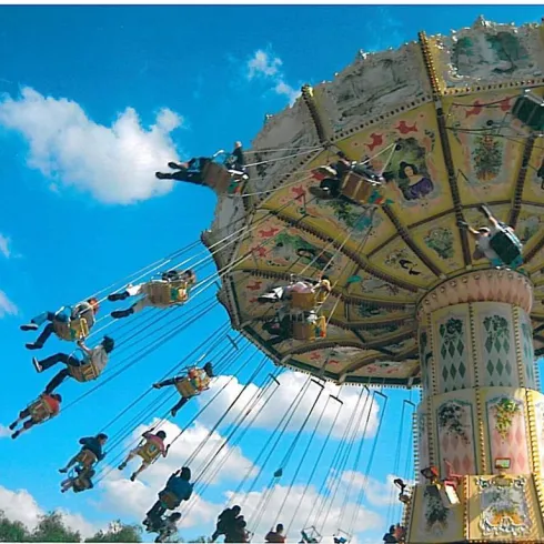 Ein bunter Freizeitpark mit einem Schwungkarussell, auf dem viele Menschen fröhlich schwingen. Der Himmel ist klar und blau, mit wenigen Wolken.