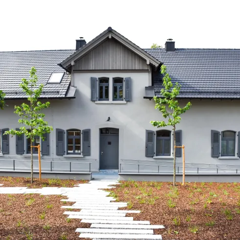 Ein modernes Haus mit grauer Fassade und einem schrägen Dach. Vor dem Haus befinden sich kleine Bäume und eine gepflasterte Auffahrt.