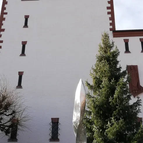 Ein historisches Gebäude mit einer spitzen Turmspitze und einem modernen, glänzenden Kunstwerk im Vordergrund. Umgeben von Bäumen und einer Steinmauer.