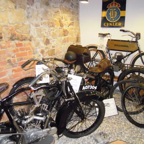 Eine Sammlung von Vintage-Fahrrädern in einem Museum. Die Fahrräder stehen vor einer bruchsteingemauerten Wand und sind mit Informationsschildern versehen.
