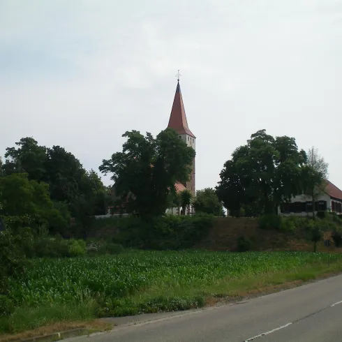 Eine Kirche mit einem roten Dach steht umgeben von Bäumen und Wiesen. Im Vordergrund verläuft eine Straße.