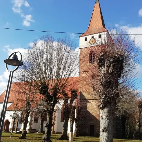 Eine alte Kirche mit einem auffälligen Turm steht in einer ruhigen Umgebung. Umgeben von beschnittenen Bäumen und einem blauen Himmel.