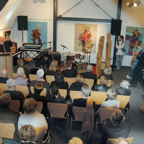 Eine Versammlung in einem Kunstraum mit einem Redner am Podium. Im Hintergrund stehen Kunstwerke und eine kleine Band.