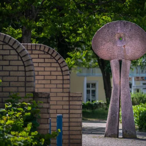 Eine moderne Skulptur steht in einem grünen Park. Im Hintergrund sind elegante Backsteinbögen zu sehen.