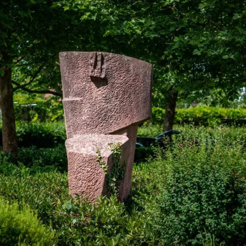 Eine abstrakte Skulptur aus rosafarbenem Stein steht in einer grünen Umgebung. Die Statue hat eine geometrische Form und ist teilweise von Pflanzen umgeben.