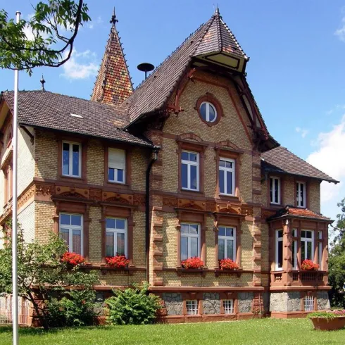 Ein charmantes historisches Haus mit spitzen Dächern und vielen Fenstern. Der Garten ist grün und mit Blumen geschmückt.