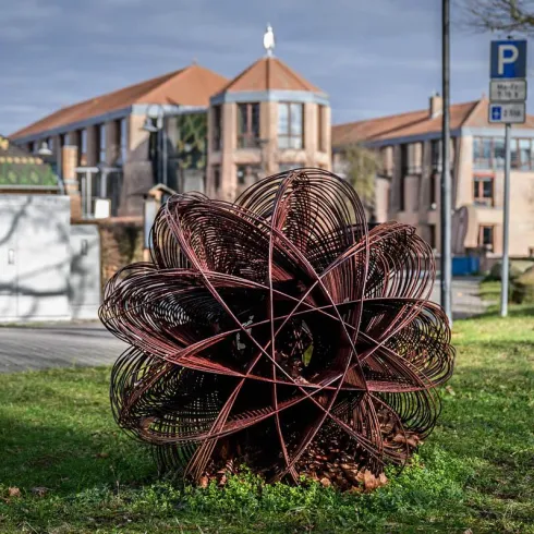 Eine kunstvolle Skulptur aus Metalldraht steht im Vordergrund. Im Hintergrund sind moderne Gebäude und ein Parkierungszeichen sichtbar.
