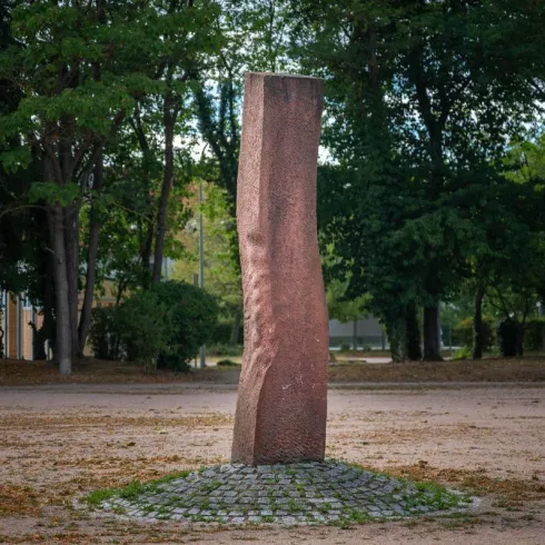 Ein hoch aufragendes, rechteckiges Denkmal aus rotem Stein steht in einem ruhigen Park. Umgeben von Bäumen und einem mit Steinen belegten Platz strahlt es eine friedliche Atmosphäre aus.
