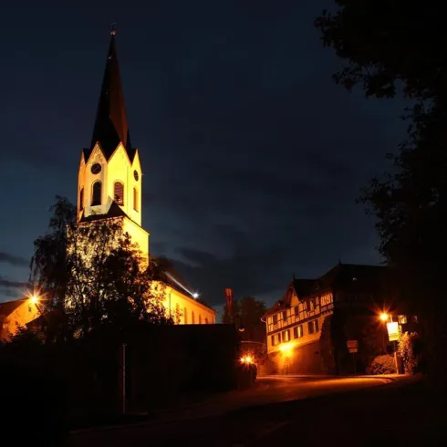 Eine schöne Kirche mit spitzem Turm, beleuchtet in der Nacht. Im Hintergrund sind alte Gebäude und Bäume sichtbar.
