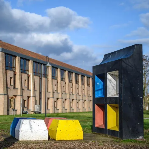 Eine moderne Skulptur mit bunten Elementen steht auf einer grünen Wiese. Im Hintergrund sieht man ein großes Bauwerk mit Fenstern und einem Dach.