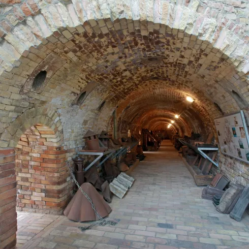 Ein alter, gewölbter Gang mit Ziegelsteinen und sanfter Beleuchtung. An den Wänden sind verschiedene Utensilien und Gegenstände ausgestellt.
