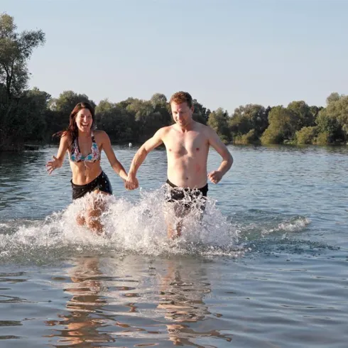 Ein Paar läuft lachend ins Wasser und spritzt dabei. Im Hintergrund sind Bäume und eine ruhige Wasseroberfläche zu sehen.