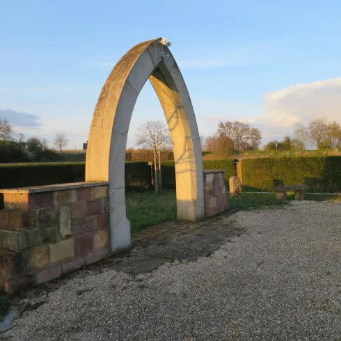 Ein eleganter Bogen aus Stein steht in einer grünen Landschaft. Im Hintergrund sind Sträucher und ein klarer Himmel sichtbar.