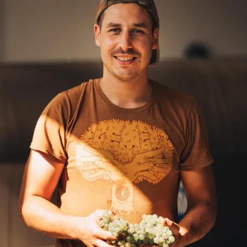 Ein junger Mann mit einem lächelnden Gesicht steht drinnen und hält Weintrauben in der Hand. Er trägt ein braunes T-Shirt und hat eine lässige Ausstrahlung.