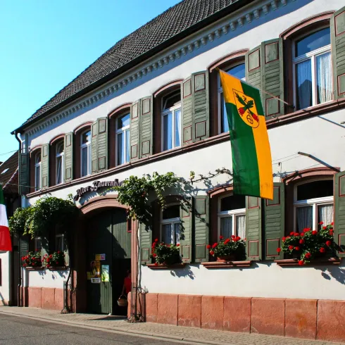 Ein traditionelles Gebäude mit grünen Fensterläden. Vor dem Gebäude hängen die Flaggen Italiens und einer Region, umgeben von blühenden Pflanzen.