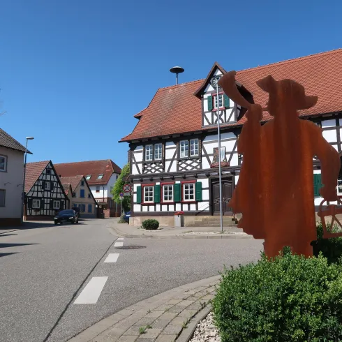 Eine malerische Straße mit traditionellen Fachwerkhäusern und klarem blauen Himmel. Im Vordergrund steht eine kunstvolle Figur.