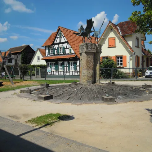Ein Spielplatz mit einer Holzplattform und einem alten Brunnen. Im Hintergrund sind charmante, traditionelle Häuser mit roten Dächern zu sehen.