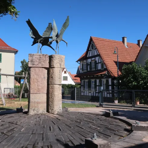 Ein Brunnen mit einer Skulptur von Vögeln steht im Mittelpunkt eines Platzes. Umgeben ist er von traditionellen Gebäuden und grünen Bäumen.