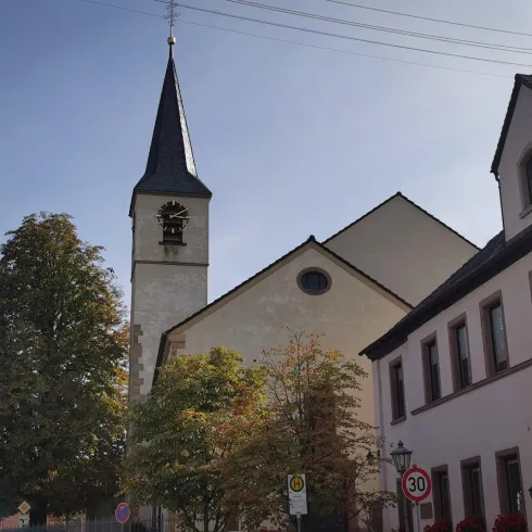 Eine schöne Kirche mit einem hohen Kirchturm steht inmitten von Bäumen. Neben der Kirche sind einige Gebäude zu sehen.