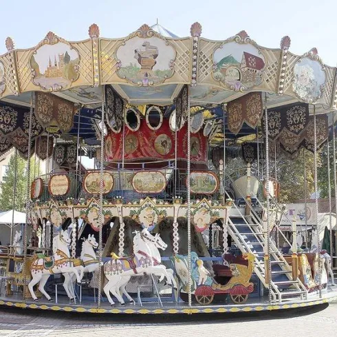 Ein nostalgisches Karussell mit bunten Figuren und dekorativen Elementen steht auf einem Platz. Es ist ein sonniger Tag und die Umgebung wirkt einladend.