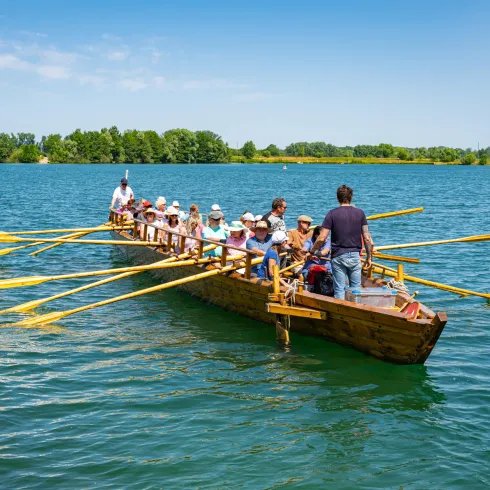 Eine Gruppe von Menschen rudert gemeinsam in einem großen Boot auf einem ruhigen Gewässer. Im Hintergrund sind grüne Bäume und ein klarer, blauer Himmel zu sehen.