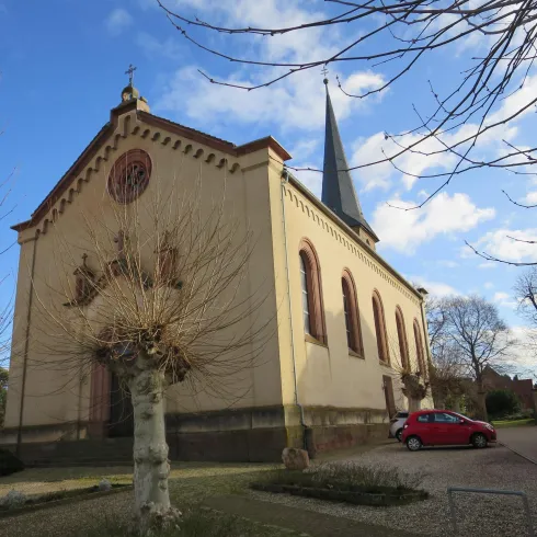 Eine Kirche mit einem spitzen Turm und großen Fenstern. Der Himmel ist teilweise bewölkt und es gibt kahle Bäume im Vordergrund.