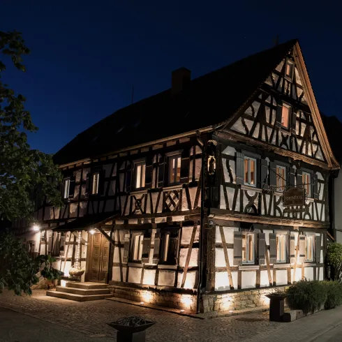 Ein historisches Fachwerkhaus bei Nacht, beleuchtet und einladend. Es hat grüne Pflanzen und einen gepflasterten Weg vor der Tür.