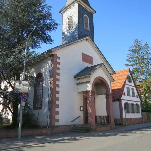 Eine kleine Kirche mit einem hohen Turm und einem bunten Dach. Die Umgebung ist von Bäumen und Gebäuden umgeben.