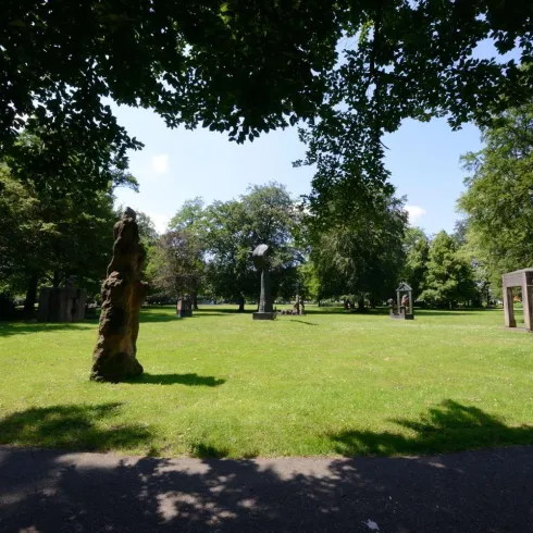 Ein sonniger Park mit grünem Rasen und mehreren Skulpturen. Umgeben von Bäumen bietet der Platz eine entspannte Atmosphäre.