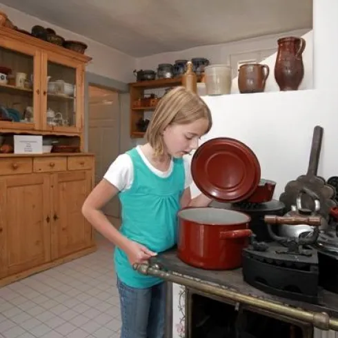 Ein Mädchen steht in einer Küche und schaut in einen roten Topf. Im Hintergrund sind verschiedene Töpfe und ein Holzschrank zu sehen.