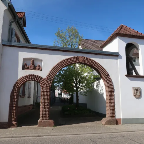 Ein historisches Tor mit einem runden Bogen und einer kleinen Uhr auf der Seite. Im Hintergrund sind Wohngebäude und ein Baum sichtbar.